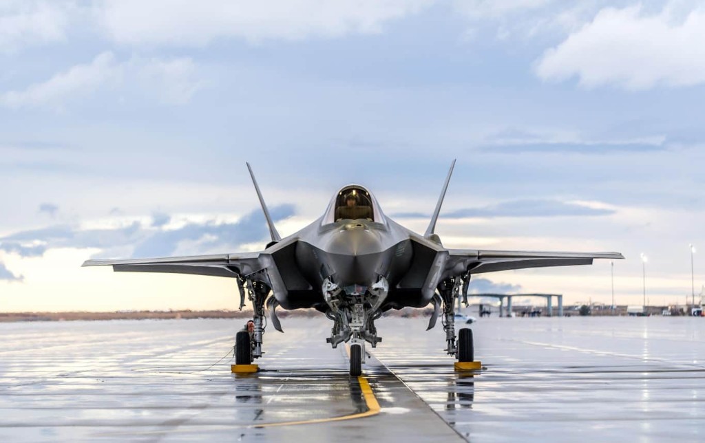 Fighter jet head-on on a wet reflective runway under a cloudy sky